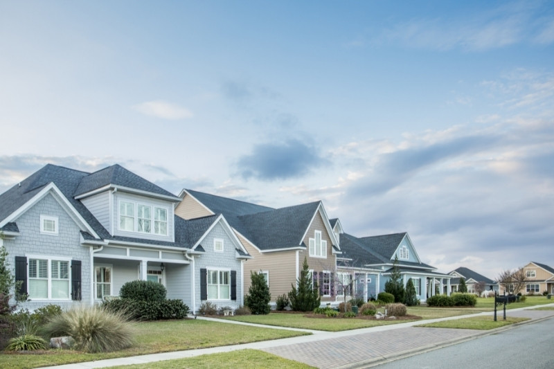 A row of modern suburban houses with well-kept lawns and landscaping under a partly cloudy sky, lining a quiet residential street.