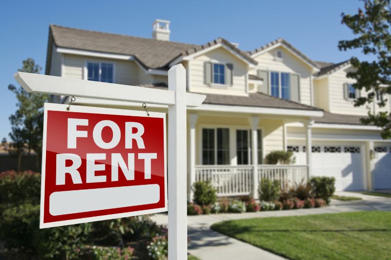 A red and white For Rent sign is posted in front of a two-story, yellow house with a white fence, porch, and well-kept yard on a sunny day.